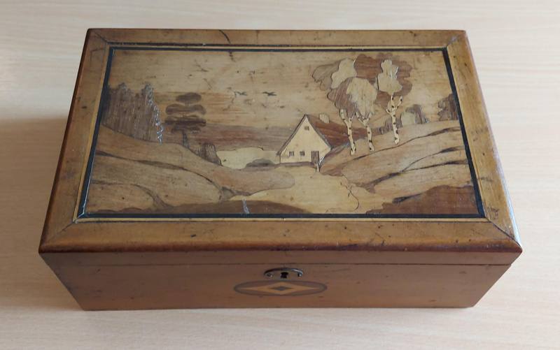 Wooden box viewed off-centre from above. The lid of the box shows a landscape scene with small white building in the middle.