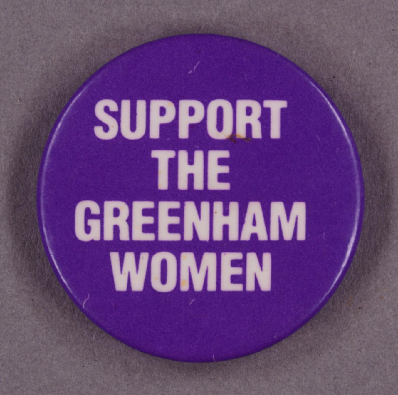 Digital image of a purple badge with white text saying: "Support The Greenham Women"